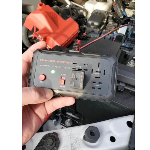 Hand holding Automotive Relay Tester Kit 12V Handheld Diagnostic Tool for 4 and 5 Pin Relays With Battery Clips in engine bay.