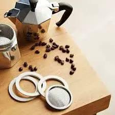 Close-up of three seals and one filter for 6-cup Bialetti Moka Express on wooden table; moka pot and coffee beans visible.