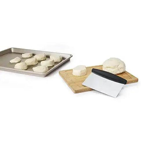 Stainless Steel All Purpose Scraper and Dough Cutter for Pizza Bread and Cookie Dough laying on cutting board beside dough on tray