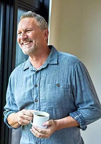 Smiling man holding a coffee mug while standing in a casual blue shirt by a window, enjoying a moment of relaxation.