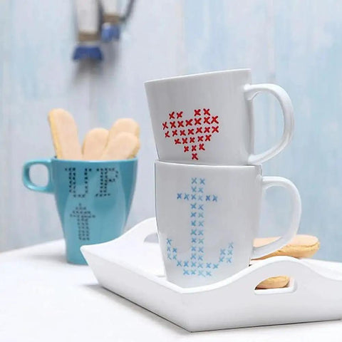 White mugs with red and blue cross-stitch patterns, stacked on a tray beside a blue mug with cookies.