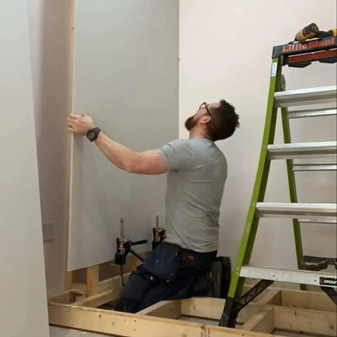Person using Original Handheld Jack and Clamp for One Hand Lifting Up to 330 Lbs to install drywall panel near a ladder