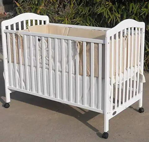 White wooden baby crib with slatted sides, beige bedding, and caster wheels, placed outdoors on concrete surface.