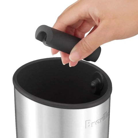 Breville BEA501BSS knock box; close-up of hand inserting a black accessory into a brushed stainless steel container.