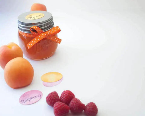 Jar of orange jam with a ribbon next to apricots and raspberries on a white surface
