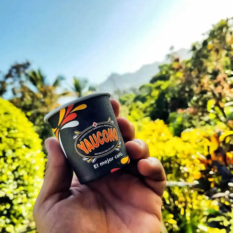 Hand holding a Yaucono coffee cup with nature and mountains in the background, vibrant and sunny setting.