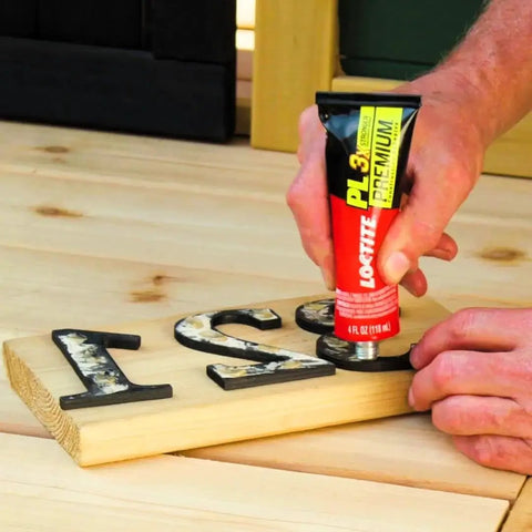 Person applying Loctite PL Premium 4 oz adhesive on wood sign indoors, product labeled as Tough As Nails Durable Polyurethane Construction Adhesive For Wood Concrete Stone And Metal 4 Fl Oz Squeeze Tube 1 Pack Repositionable Safe To Use VOC Compliant All Purpose Glue That Bonds Tight And Lasts As Long As The Surface Loctite PL Premium.