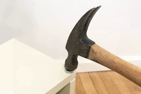 Hammer with a wooden handle removing a nail from a white piece of furniture on a wooden floor.