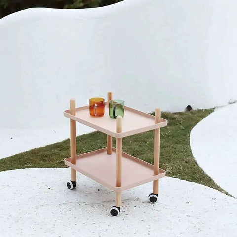 Pink two-tier cart with wooden frame and caster wheels holding two colorful cups on a minimalist white and green background.