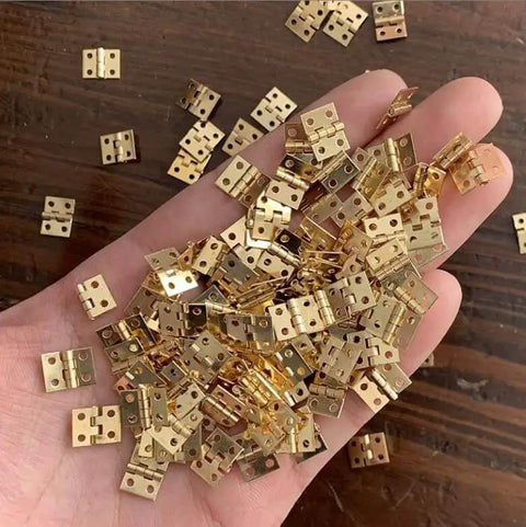 Hand holding 100 Piece Set Tiny Metal Mini Hinges With Screws For Dollhouse Furniture Cabinets Closets And Miniature Boxes Retro Style For Diy Craft Projects And Small Home Decor.