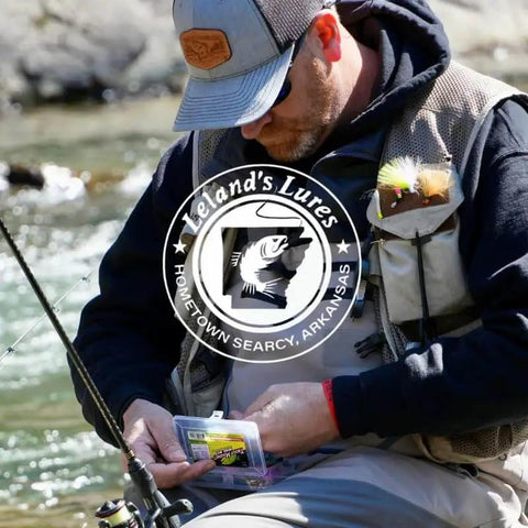 Angler holding Leland's Lures package near a river, wearing fishing vest and cap.