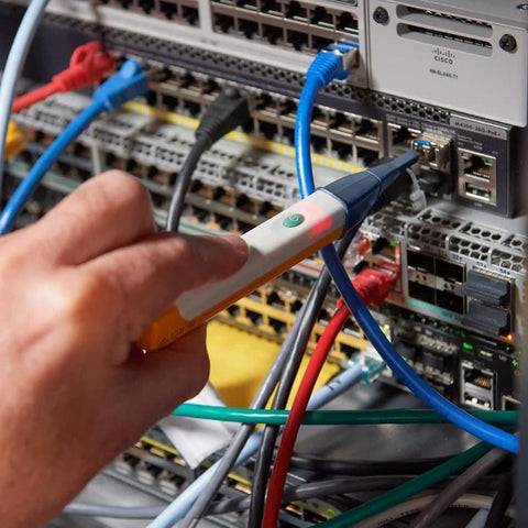 Fiber optic cable tester in hand, orange and white handheld device with green power button, connected to a blue fiber cable on a network rack.