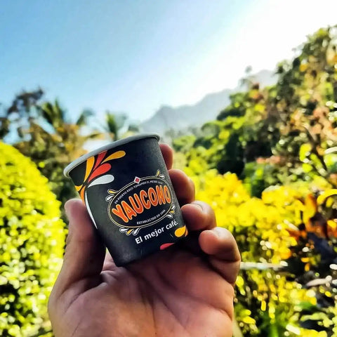 A hand holding a Yaucono espresso cup in a tropical outdoor setting with lush greenery and mountains.