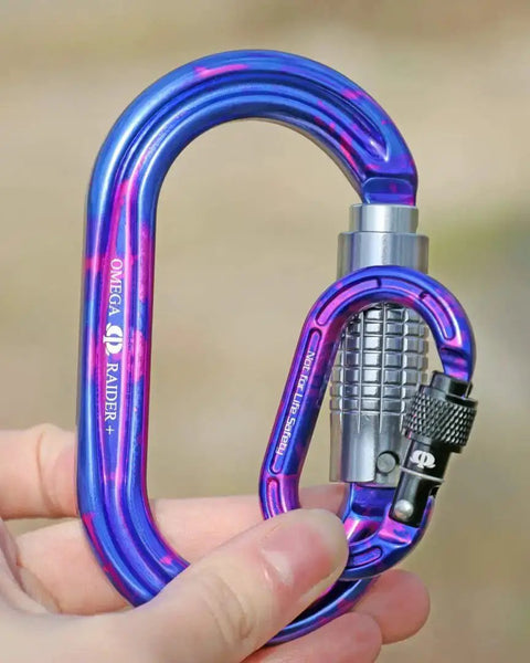 Person holding blue Omega Raider screw-lock carabiner with black locking mechanism, outdoor setting.