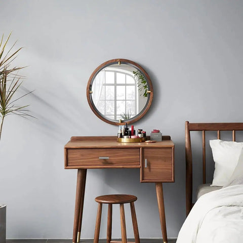 Modern wooden vanity with round mirror and stool, next to a bed in a minimalist bedroom, against grey wall.