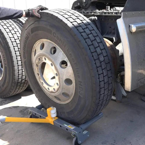 Side view of Winntec rolling wheel dolly under a large truck tire; gray steel frame with yellow handle.
