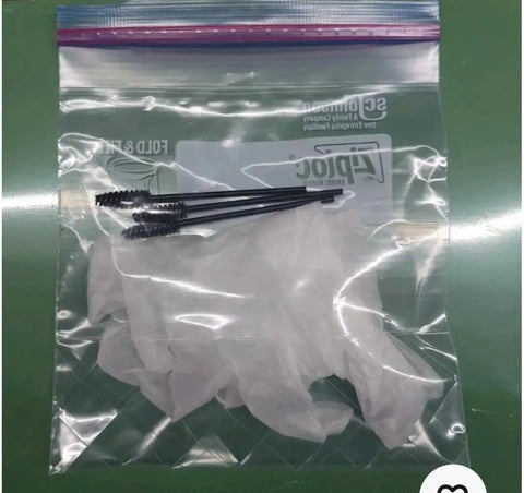 Clear plastic ziplock bag containing three eyelash brushes and a pair of white gloves on a green surface.