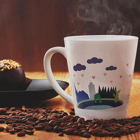 White ceramic mug with skyline and blue cloud design, surrounded by coffee beans, with a chocolate dessert in the background.