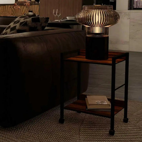 Two Tier Rolling Utility Cart With Brown Wood Shelves And Black Metal Frame On Wheels beside a sofa with decorative lamp on top