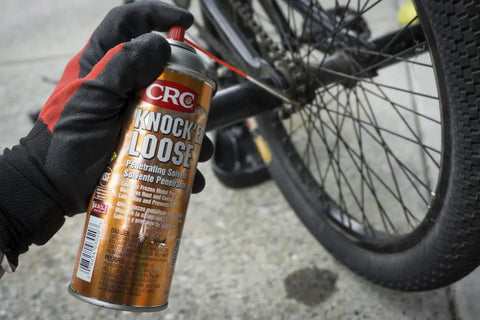 Front view of CRC Knock’er Loose penetrating solvent aerosol can, 13 oz, held by a gloved hand beside a bicycle wheel.