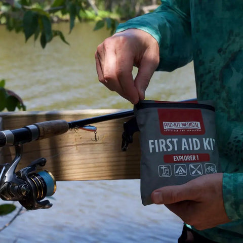 Person holding Watertight Explorer 67-Piece Outdoor First Aid Kit for Hiking and Camping near a fishing rod by a lake.