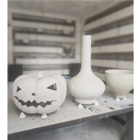 Three white ceramic pottery pieces on a shelf: carved jack-o’-lantern pumpkin, tall-neck vase, and footed bowl.