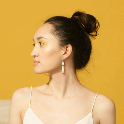 Side profile of a person with updo hairstyle wearing colorful dangle earrings against a yellow background.