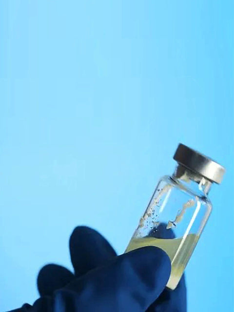 Close-up of a 15ml glass vial with a sealed metal cap held by a gloved hand against a blue background