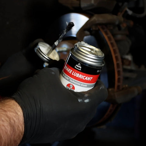 4 oz SIL-Glyde brake grease can with red/black label, brush-top, held by gloved hand near a brake caliper