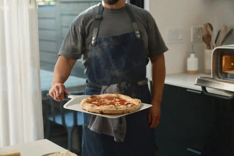 Compact 12 Inch Aluminum Pizza Peel With Handy Handle For Easy Pizza Turning And Oven Prep Lightweight Yet Durable Ideal For Home Kitchens And Ooni Ovens Space Saving Design For Quick Pies in use by chef holding a fresh pizza.