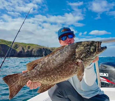 Angler holding a large fish on a boat near a shoreline, showcasing use of Offshore Fishing Speed Clip Bundle With 50 Heavy-Duty Stainless Steel Lure Clips.