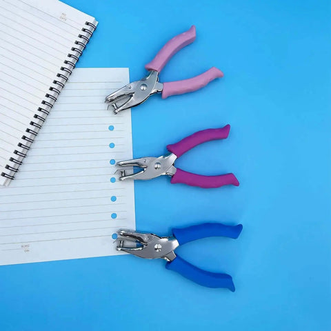Three 1/4 Inch Hole Punchers for Crafts with soft grips in pink, purple, and blue, next to punched notebook paper on a blue background.