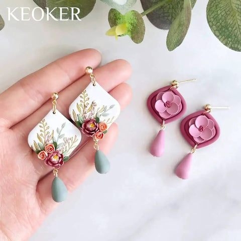 Hand holding floral polymer clay earrings with teardrop details, pink and green tones, displayed on a neutral background.