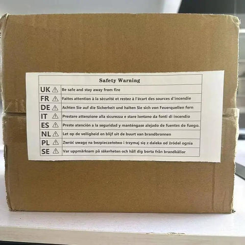 Safety warning label on a cardboard box in multiple languages, cautioning to stay away from fire, photographed on a desk.
