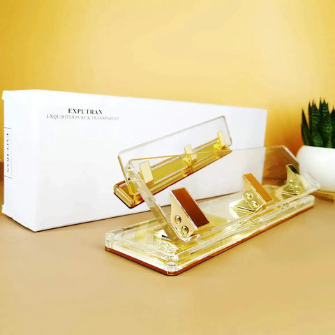 Clear Acrylic 3 Hole Punch For 10 Sheets With Four And A Quarter Inch Hole Spacing Gold Accents on desk with box and plant
