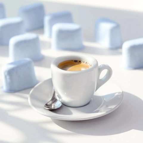 White ceramic espresso cup with saucer and spoon on a white surface; blue sugar cubes blurred in background.