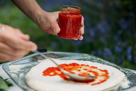 Person spreading tomato sauce on pizza dough, outdoor setting.