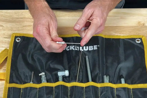Hands holding grease injector needle for sealed bearings with 18 gauge steel and protective plastic cover near LockNLube coupler case.