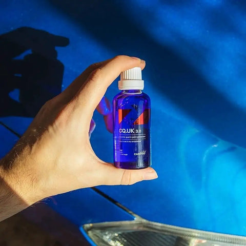 Hand holding CQUARTZ UK 3.0 Ceramic Coating Kit bottle in front of blue car paint and headlight