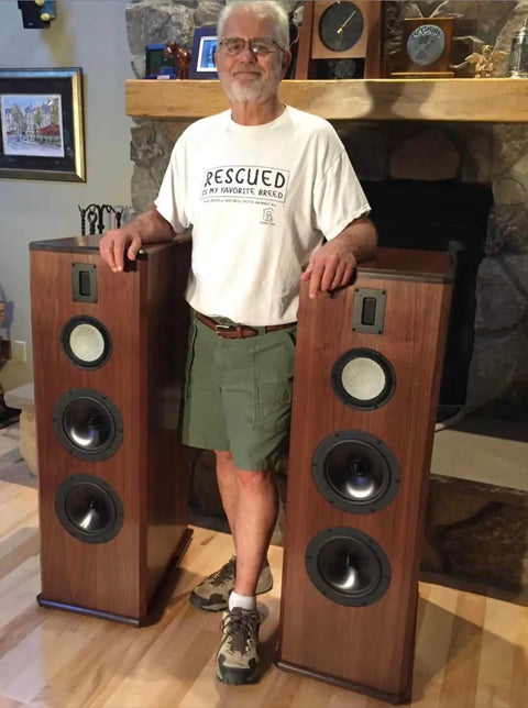 Man standing between two tall brown speakers in a living room with stone fireplace and wooden mantle.