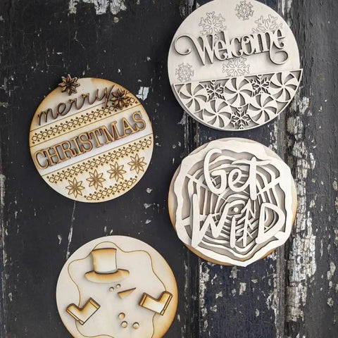 Wooden discs with laser-engraved designs including "Merry Christmas," "Welcome," "Get Wild," and a snowman, on a dark wooden background.