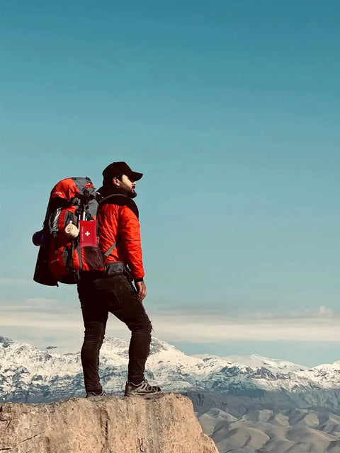 Hiker with backpack carrying Compact 97 Piece Travel First Aid Kit for Cars Home School Sports Camping and Hiking on a mountain top