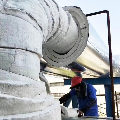 White ceramic fiber blanket insulation roll wrapped around a large metal pipe; worker in blue jacket and red hard hat.