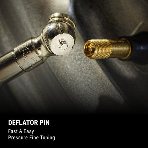 Pencil-style tire pressure gauge with built-in deflator pin, plated brass finish, front view beside a tire valve.