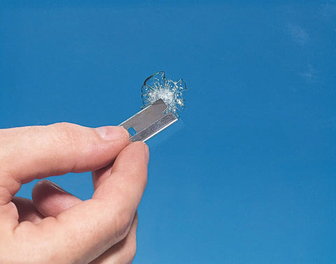 Close-up of a hand holding a razor blade with a chip repair patch from Windshield Repair Kit From Rain X For Chips Cracks Bulls Eyes Stars And Laminated Glass With Advanced Resin Formula Color Clear Great For Multiple Repairs.