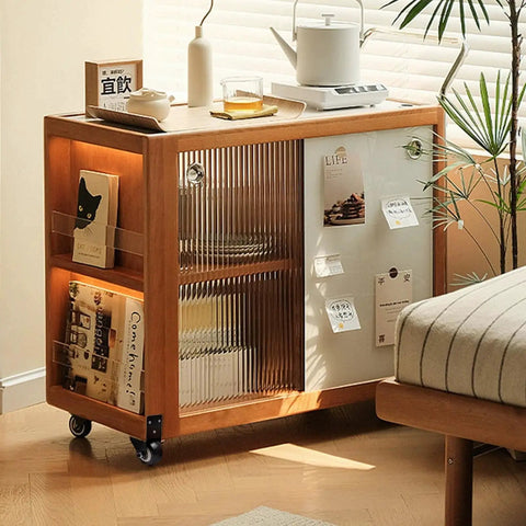 Wooden storage cabinet on wheels next to a bed with books, decorative items, and glass sliding door in a sunlit room.