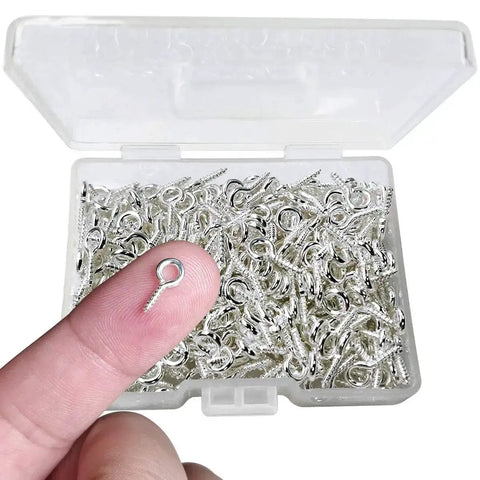 Close-up of a finger holding a silver screw eye pin over a box of 300Pcs Mini Screw Eye Pins and Hooks Set 10x5mm SelfTapping Screws for Arts and Crafts DIY Jewelry Making Cork Top Bottles and Christmas Ornaments Silver Super Handy for DIYers