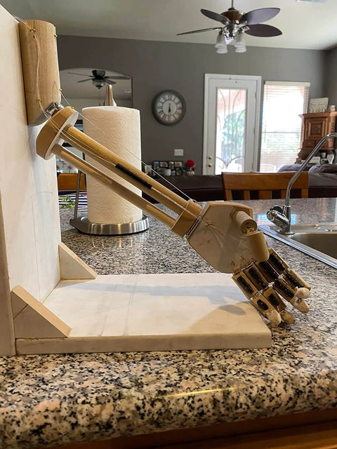 Wooden mechanical arm model extending towards countertop in a kitchen setting, near paper towel holder and kitchen sink.