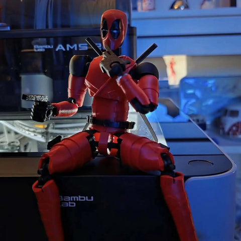 Red and black action figure sitting on a table with blue background, near a Bambu Lab AMS 3D printer.