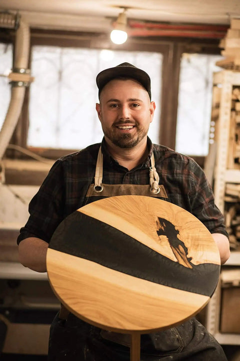 Woodworker holding a round wooden tabletop with black stripe and silhouette design in a workshop setting.